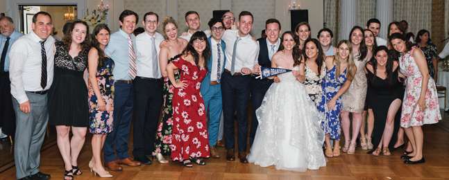 Large group of people pose around a woman in a white wedding dress who's holding a small Brandeis pennant.